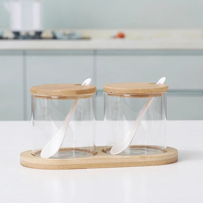 Two empty cylindrical glass jars with wooden lids and spoons, on a bamboo tray.