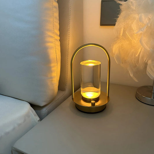 A lit, modern, portable table lamp with a gold metal frame and a glass shade, sitting on a bedside table next to a bed.