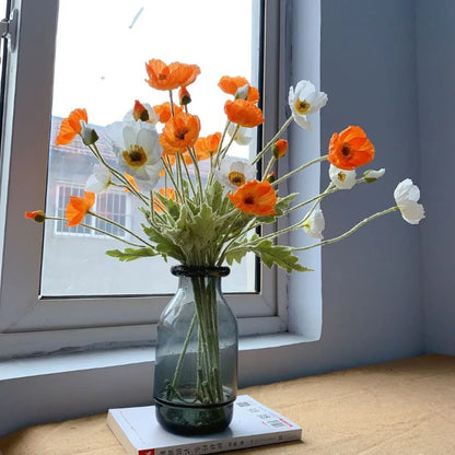 A bouquet of orange and white poppies sits in a gray vase on a book, in front of a window.