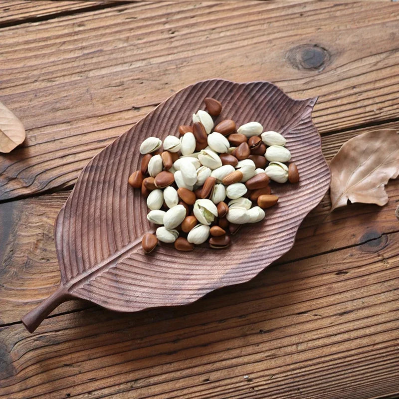 A wooden, veined leaf-shaped platter holding a mix of nuts, including pine nuts and pistachios, on a wooden table.