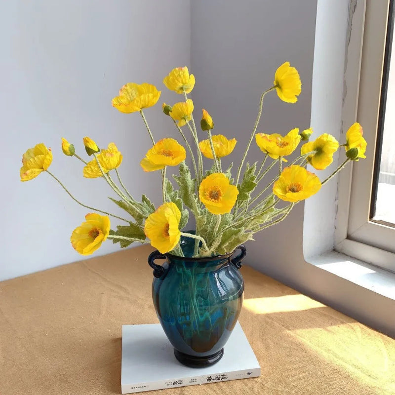 A turquoise vase holds a bouquet of yellow poppies, sitting on a white book.