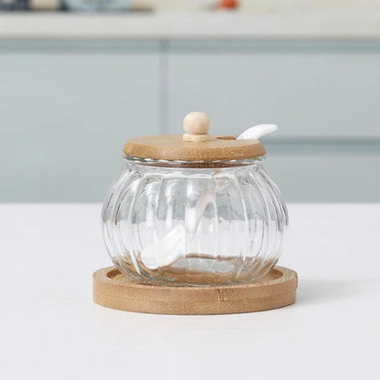 An empty pumpkin-shaped glass jar with a wooden lid, spoon, and bamboo coaster.