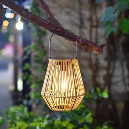 A warm-toned, woven outdoor lantern with vertical slats and a dark solar panel on top hangs from a branch at night, emitting a soft glow. The lantern has a metal handle. Blurred lights and greenery are visible in the background.