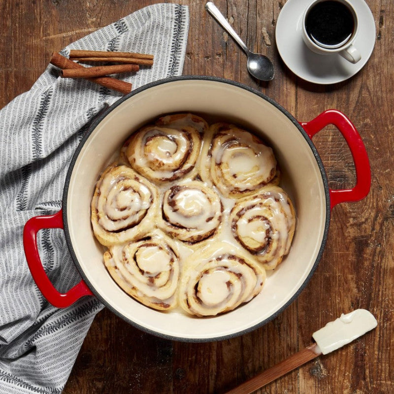 Cinnamon rolls in a red baking dish on a wooden table with coffee and cinnamon sticks.