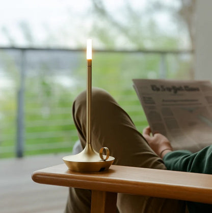 Person reading a newspaper with a gold lamp on a table by a window