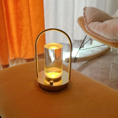
A lit, portable table lamp with a gold metal frame and a glass shade, placed on an orange upholstered surface, with an orange curtain and a rocking chair in the background.