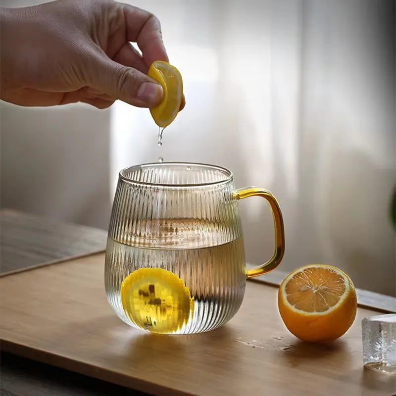 A hand squeezing a lemon wedge into a ribbed glass mug with a gold handle, which already contains water and a lemon slice. Another lemon half and an ice cube sit on a tray next to the mug.