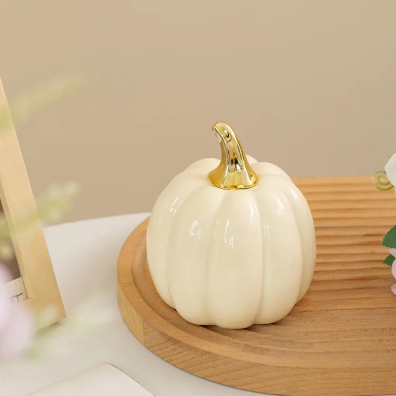 A single cream-colored ceramic pumpkin with a gold stem sits on a wooden tray, with a blurred framed picture and floral arrangement in the background.