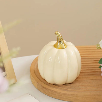 A single cream-colored ceramic pumpkin with a gold stem sits on a wooden tray, with a blurred framed picture and floral arrangement in the background.