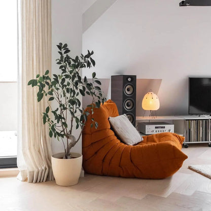 An orange Togo chair with a white pillow next to a potted plant and a speaker.