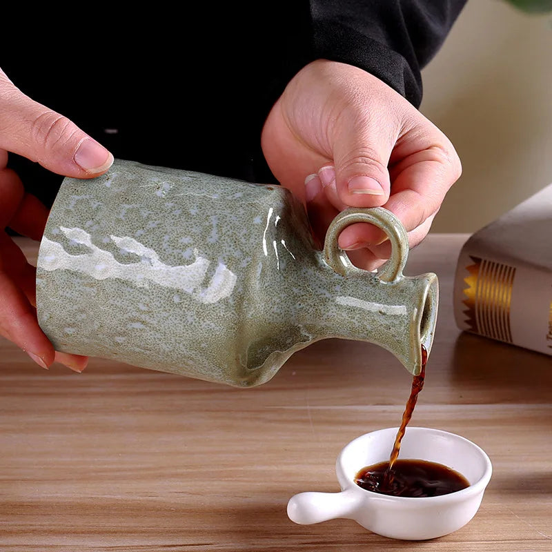 Person pouring coffee from a green ceramic pitcher into a small white cup on a wooden surface.