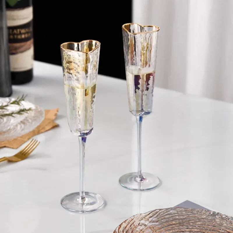 Two elegant heart-shaped champagne flutes with gold rims on a white marble table.