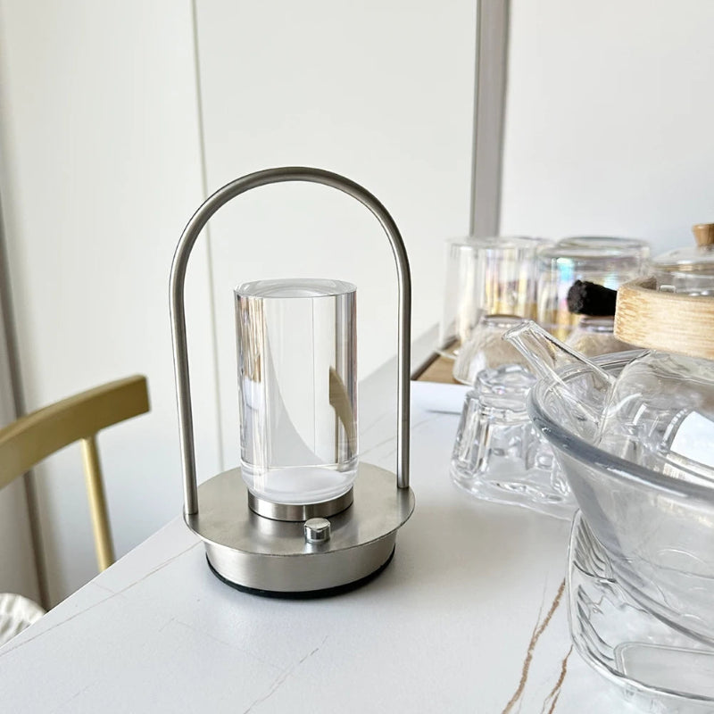 A portable table lamp with a silver metal frame and a glass shade, placed on a white table with other glassware in the background.