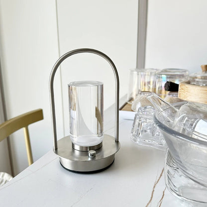 A portable table lamp with a silver metal frame and a glass shade, placed on a white table with other glassware in the background.