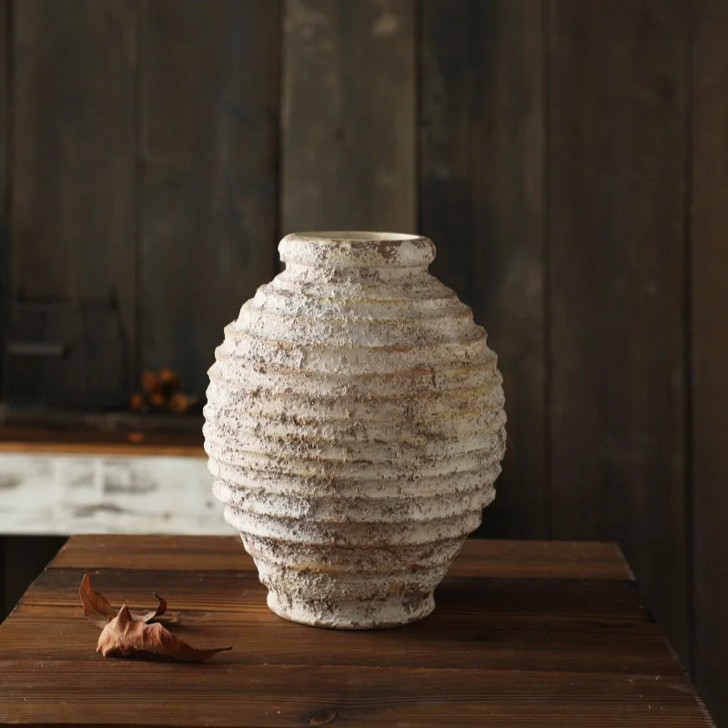 A rustic, round ceramic vase with a heavily textured, white and brown ribbed surface sits on a dark wooden table.