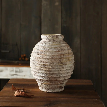 A rustic, round ceramic vase with a heavily textured, white and brown ribbed surface sits on a dark wooden table.
