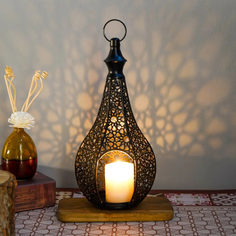 A single black ornate lantern with a lit candle sits on a wooden base, casting a patterned shadow on the wall, with a decorative vase to its left.