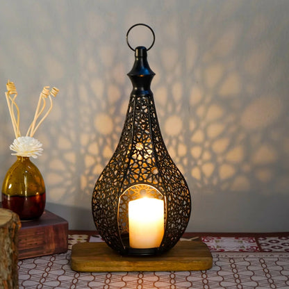 A single black ornate lantern with a lit candle sits on a wooden base, casting a patterned shadow on the wall, with a decorative vase to its left.