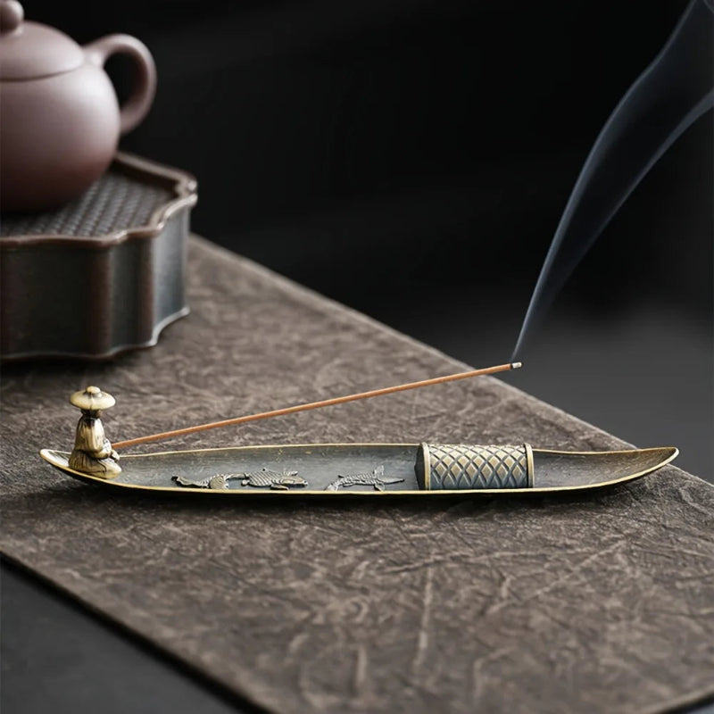 Decorative incense holder with incense sticks on a textured surface, with a blurred teapot in the background.