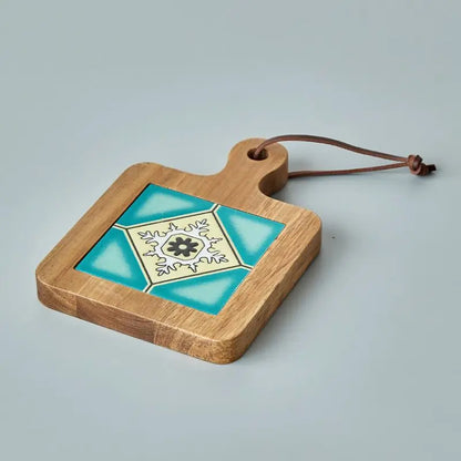 A wooden trivet with a ceramic tile inlay featuring a geometric pattern in shades of blue, white, and black rests on a light gray surface. The trivet has a handle with a hole and a leather loop for hanging.