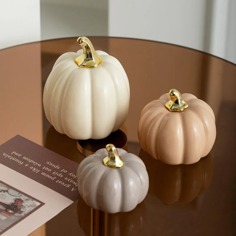 Three ceramic pumpkins in white, beige, and light gray with gold stems sit on a reflective brown surface next to a book.