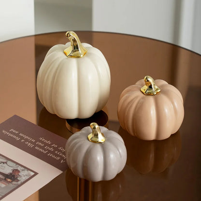 Three ceramic pumpkins in white, beige, and light gray with gold stems sit on a reflective brown surface next to a book.
