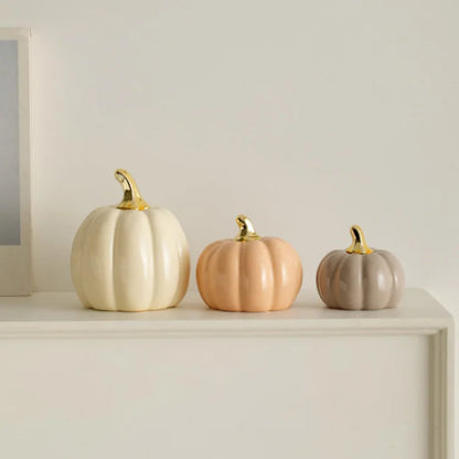 Three ceramic pumpkins of varying sizes and colors (cream, peach, and gray) with gold stems are lined up on a white mantelpiece next to a partial picture frame.