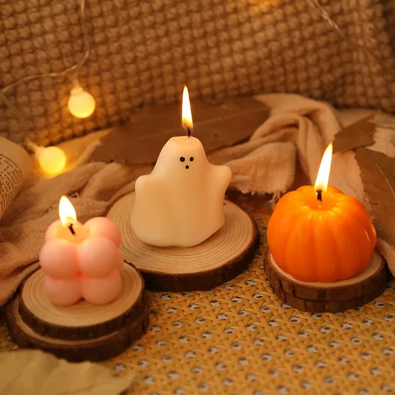 Three lit Halloween-themed candles sit on small wooden slices. From left to right, there is a light pink four-lobed candle, a white ghost-shaped candle with black dot eyes, and an orange pumpkin-shaped candle with carved vertical lines. The candles are surrounded by scattered artificial autumn leaves and warm-toned string lights in the blurred background.