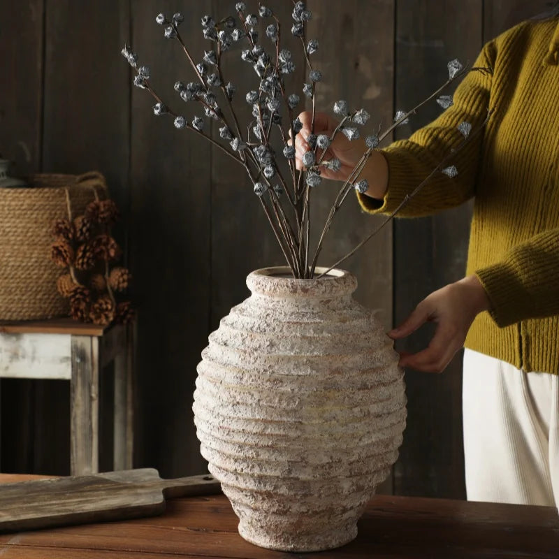 A person in a yellow sweater arranges dried blue-grey seed pods in a large, textured, rustic ceramic vase on a dark wooden table.