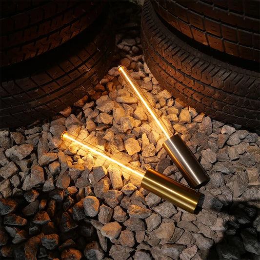 Two cylindrical lamps with glowing tubes lying on a pile of rocks with large tires in the background. One lamp has a gold base, the other has a dark bronze base.