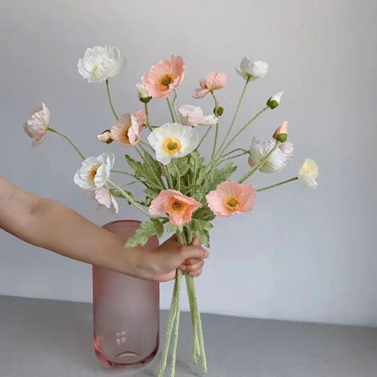 A hand holds a bouquet of white and pink poppies above a pink vase.