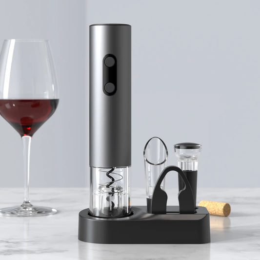 A gray electric wine opener stands on a black charging base on a light-colored surface. A clear wine glass filled with red wine is visible on the left, and a wine cork is on the right. The wine opener set includes a wine pourer and two vacuum wine stoppers. The background is a soft, light color.