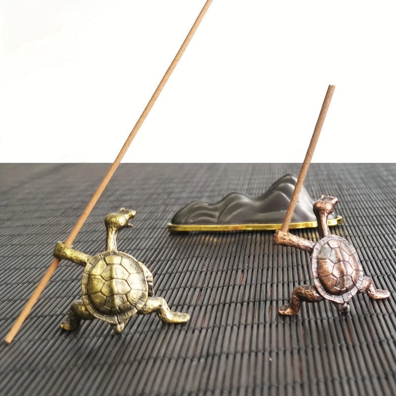 Two different colored turtle-shaped incense holders, one brass and one bronze, holding incense sticks on a dark woven mat.