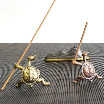 Two different colored turtle-shaped incense holders, one brass and one bronze, holding incense sticks on a dark woven mat.