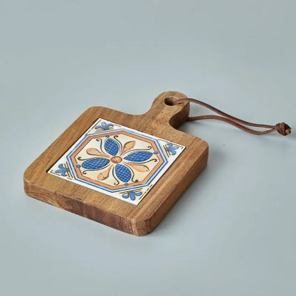 A wooden trivet with a ceramic tile inlay featuring a symmetrical floral design in blue, orange, and white rests on a light gray surface. The trivet has a handle with a hole and a brown leather loop for hanging.