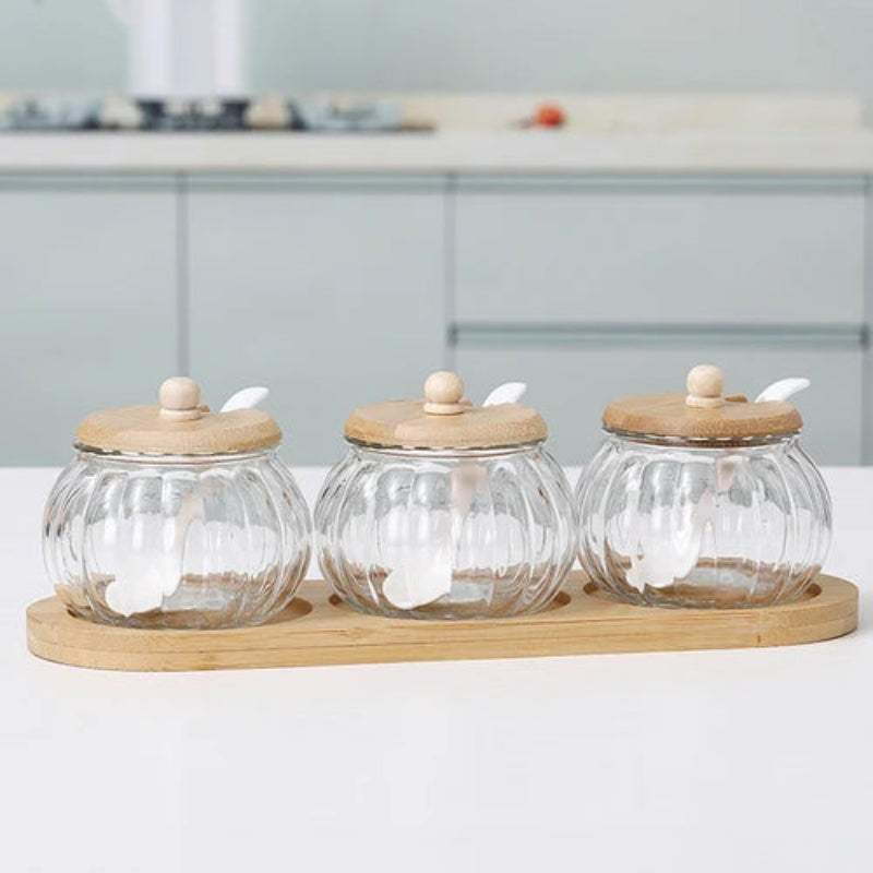 Three empty pumpkin-shaped glass jars with wooden lids and spoons, on a bamboo tray.