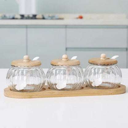 Three empty pumpkin-shaped glass jars with wooden lids and spoons, on a bamboo tray.