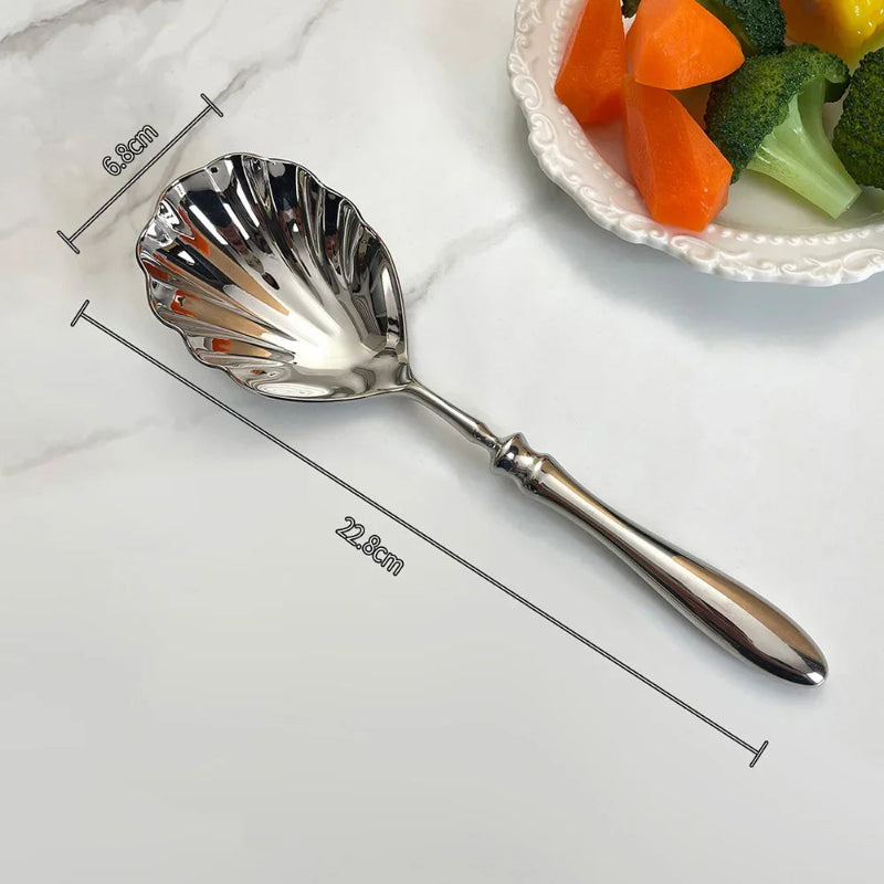 A single, ornate silver serving spoon with a scalloped, shell-shaped head, lying on a marble surface. The image is labeled with its dimensions.