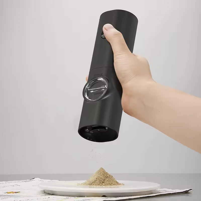 A hand holding and operating a black automatic spice grinder, with a pile of ground spices on a plate below.