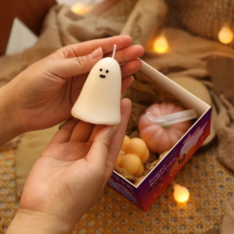Two hands gently hold a small, white, ghost-shaped candle with two black dot eyes and a curved mouth. The candle has a white wick extending from the top. Below, a partially visible open box contains a pink pumpkin-shaped candle and two stacked orange cube-shaped candles. Soft, warm-toned string lights are blurred in the background. The hands appear to be light-skinned.