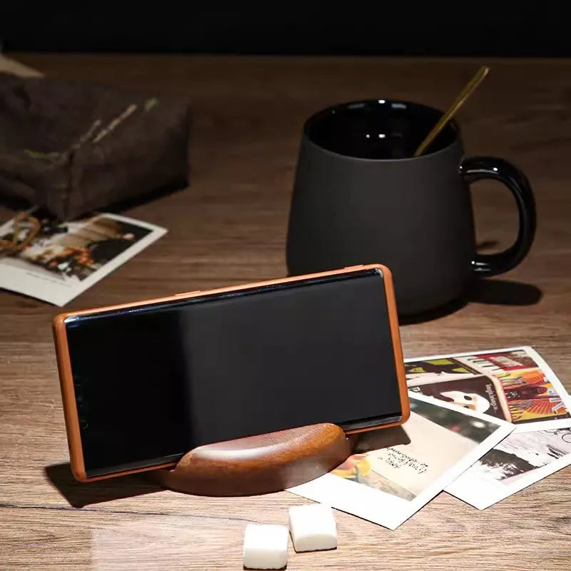 A brown smartphone in a wooden stand, a black mug with a spoon, and two sugar cubes on a wooden table with scattered photographs.