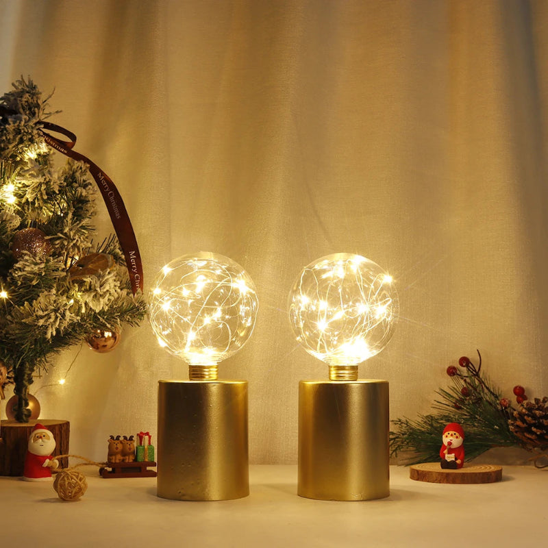 A pair of modern table lamps with gold cylindrical bases and clear glass globe shades filled with warm white fairy lights, with a miniature decorated Christmas tree and small decorations in the background.