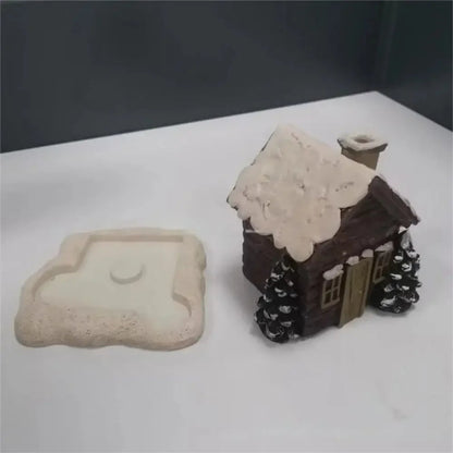 Small gingerbread house with snow on the roof and two trees on a white surface.