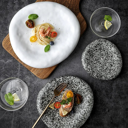 Two plates of food on a dark surface with glasses and a cutting board.