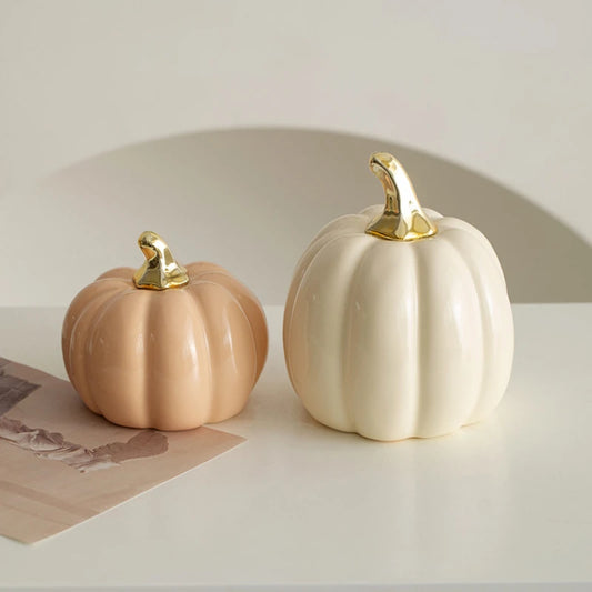 Two ceramic pumpkins, one small and peach-colored, the other larger and cream-colored, both with gold stems, sit on a white surface with a decorative arched background.