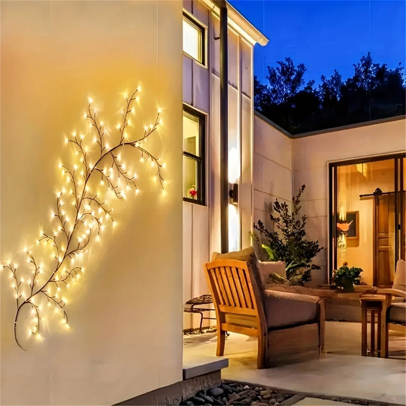 A string of lit, branch-shaped lights hangs on a white wall in an outdoor patio area with wooden furniture and a house in the background.