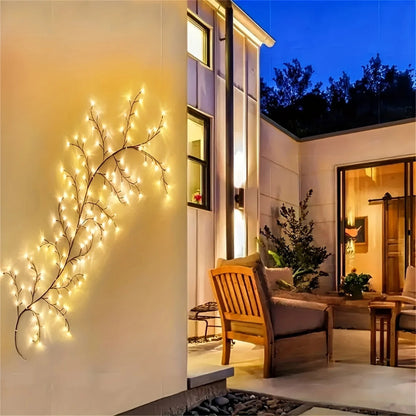 A string of lit, branch-shaped lights hangs on a white wall in an outdoor patio area with wooden furniture and a house in the background.