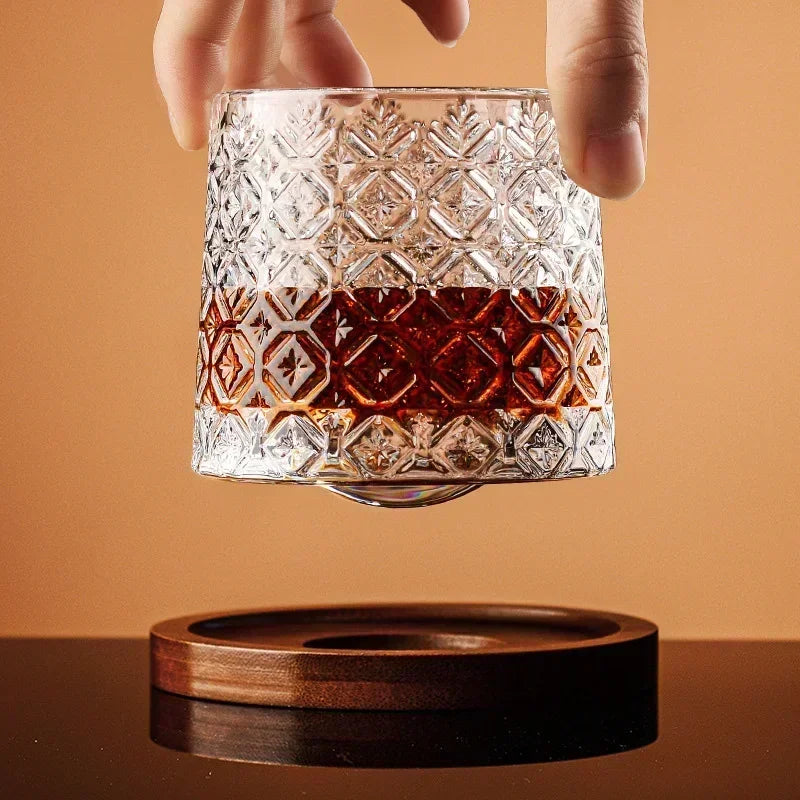 A hand holds a cut-crystal whiskey glass filled with a brown liquid, suspended just above a wooden coaster.
