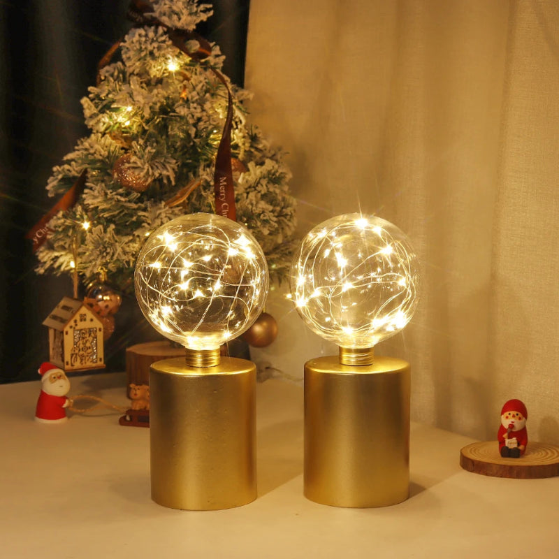 A pair of modern table lamps with gold cylindrical bases and clear glass globe shades filled with warm white fairy lights, with a miniature Christmas tree and small decorations in the background.