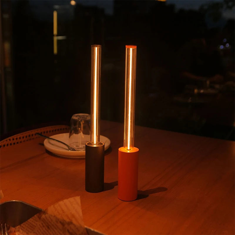 Two long, cylindrical lamps, one black and one red, with a gold tube on top that glows with warm light, sitting on a wooden table.